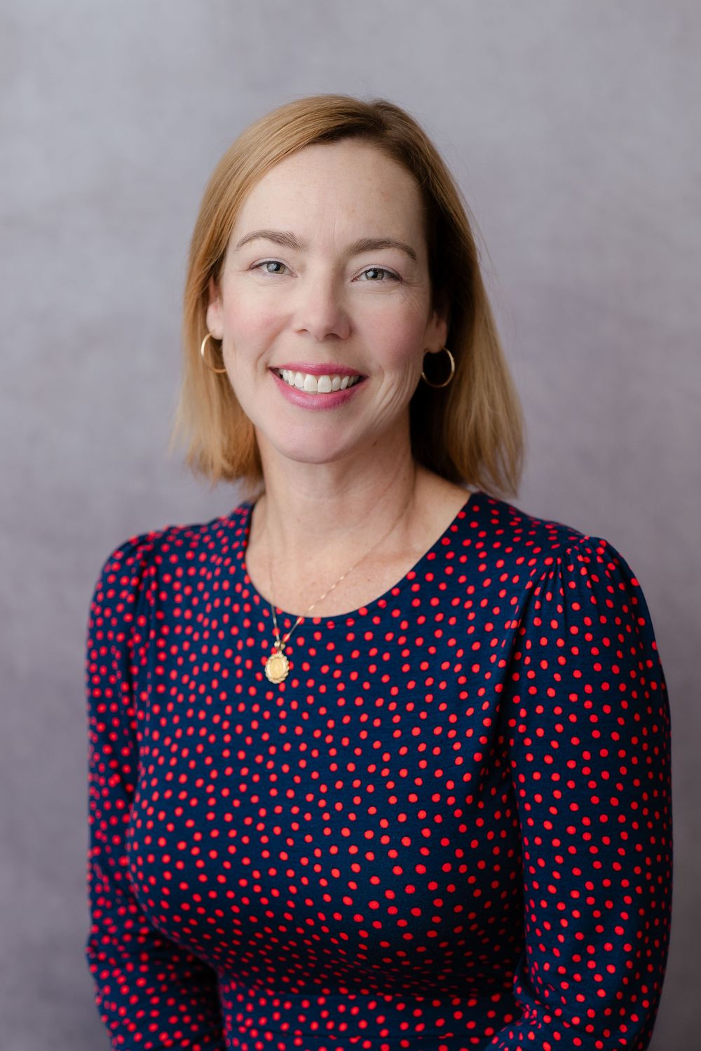 Smiling woman with red hair wearing a navy blue polka dot dress and gold necklace against a gray background.