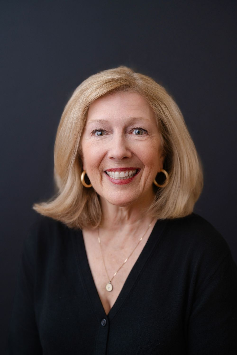 Smiling blonde woman wearing gold hoop earrings, gold necklace, and black top against dark background.