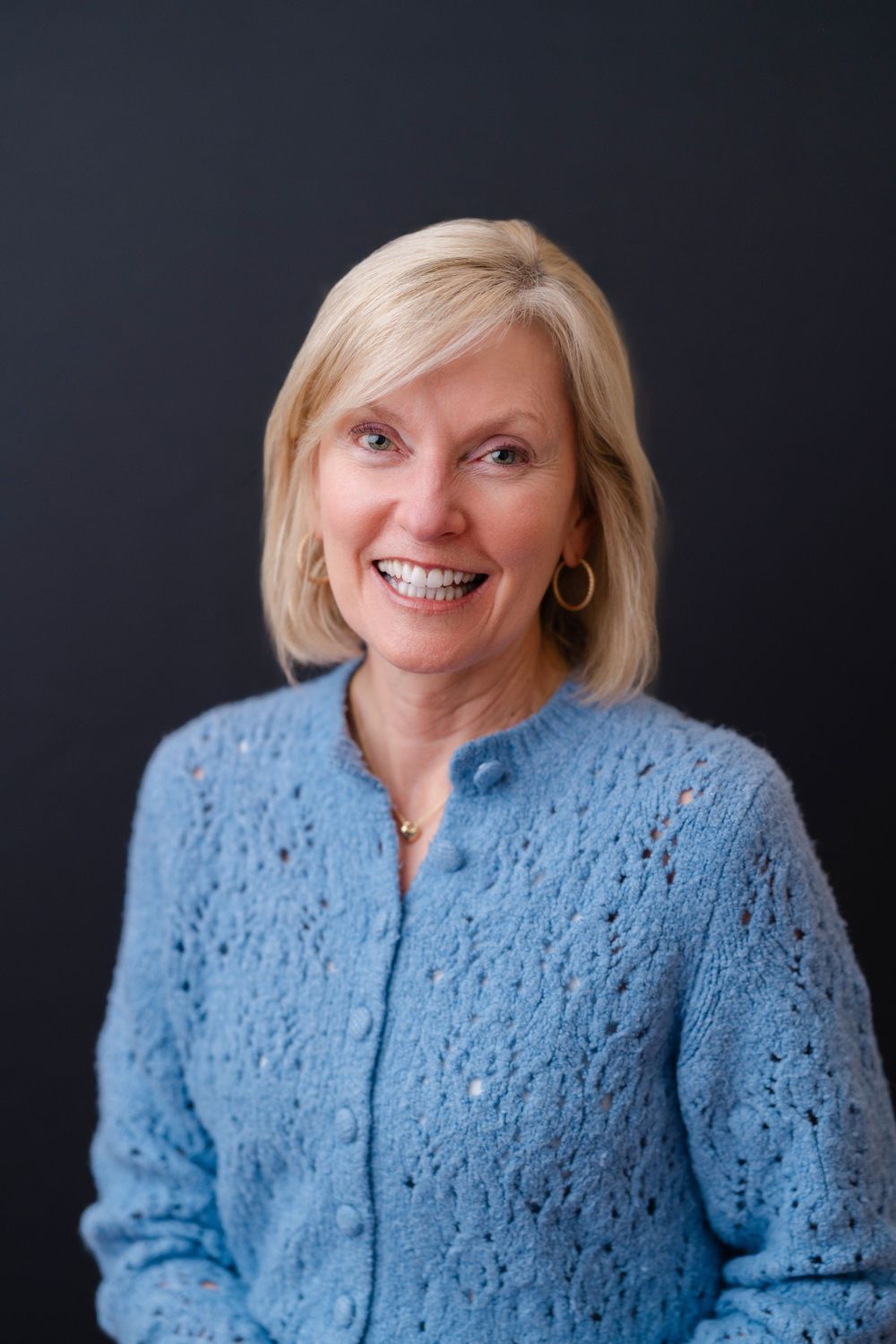 Smiling blonde woman in a blue knit cardigan posing against a dark background for a professional portrait.