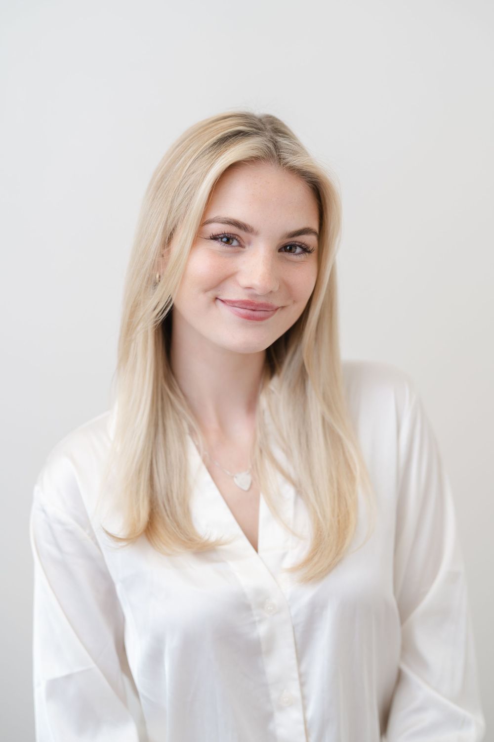 Young blonde woman smiling in white satin blouse with delicate necklace against light background.