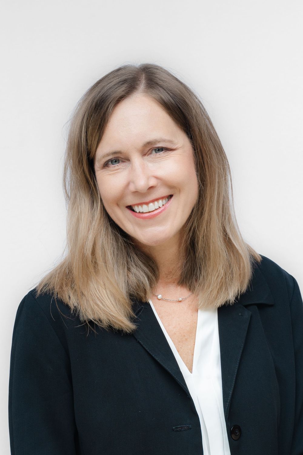 Professional headshot of a smiling woman with blonde hair wearing a dark blazer and white blouse against a light background.