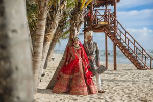 Traditional Indian wedding couple in red lehenga and grey suit posing near beach stairs.