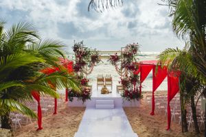 Elegant beach wedding setup with white aisle runner, floral arch, and red decorative elements.