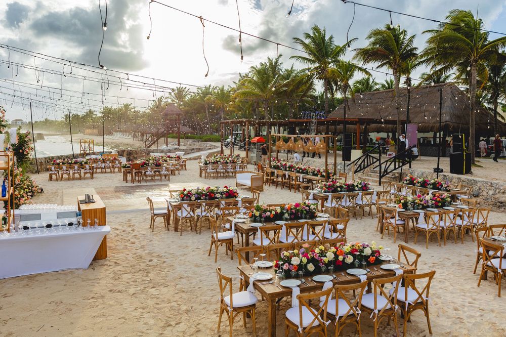 Beach reception setup with wooden chairs and tables under string lights and palm trees.