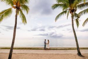 Scenic beach view with two silhouettes between tall palm trees during a cloudy evening.