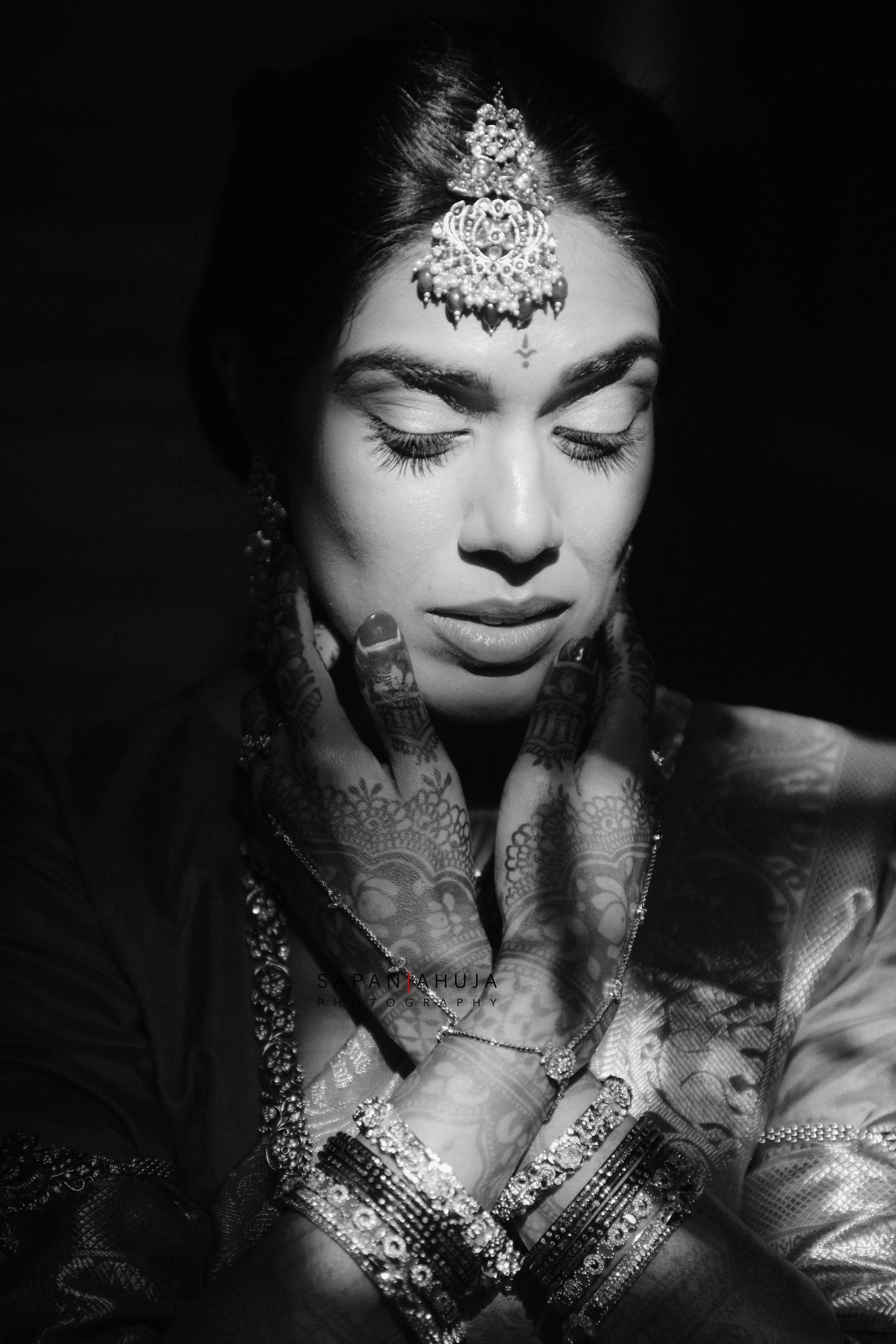 Black and white portrait of a bride with mehndi hands, ornate maang tikka jewelry, and closed eyes in prayer pose.