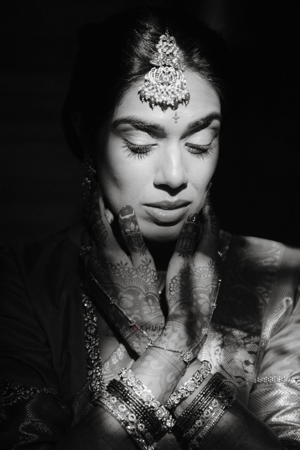Black and white portrait of a bride with mehndi hands, ornate maang tikka jewelry, and closed eyes in prayer pose.
