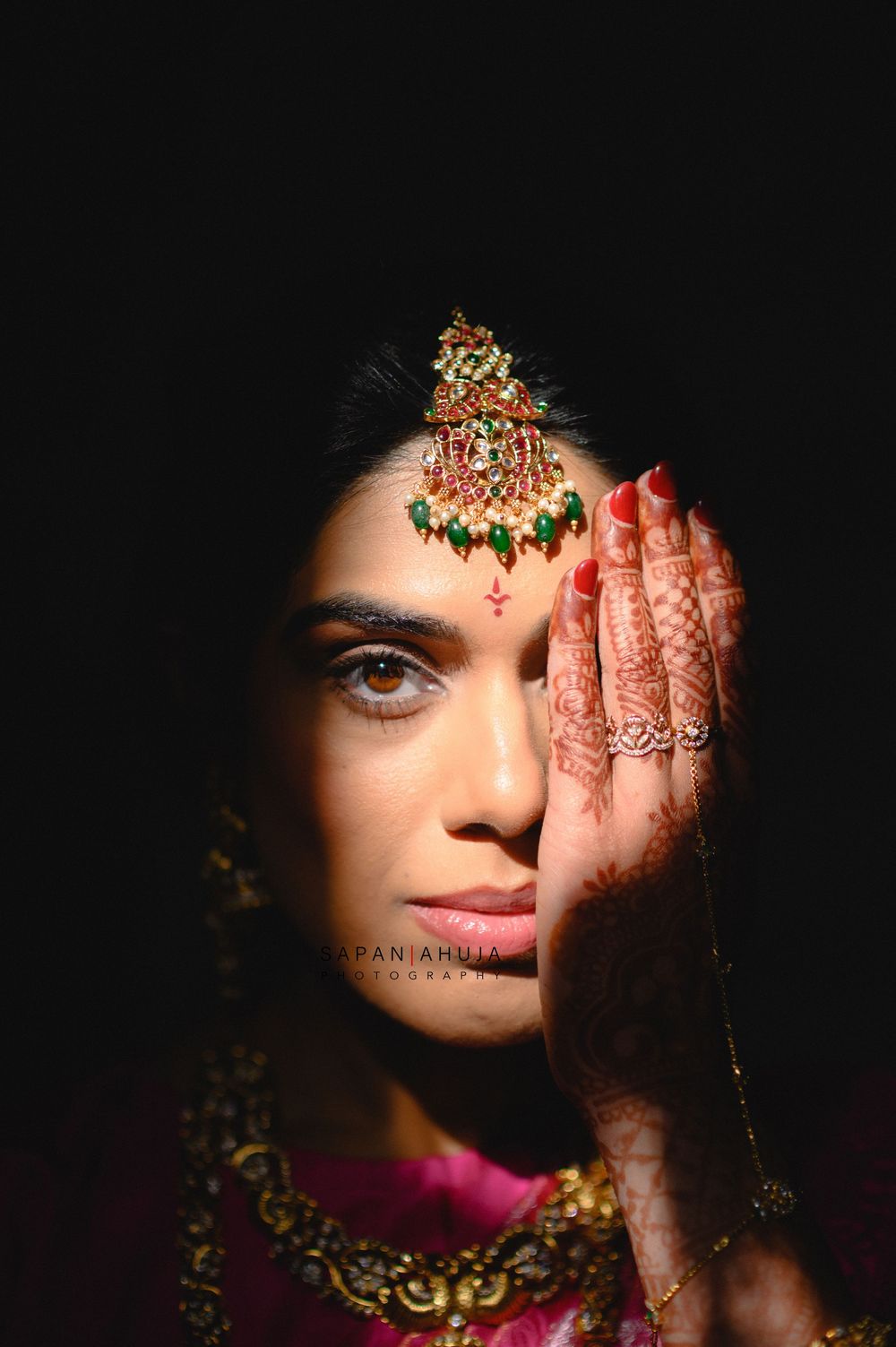 Indian bride with mehndi-covered hand holding ornate maang tikka jewelry near her face against dark background.