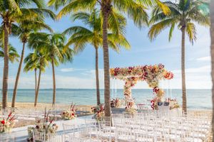 Beachfront wedding ceremony setup with pink floral arch and white chairs under palm trees.