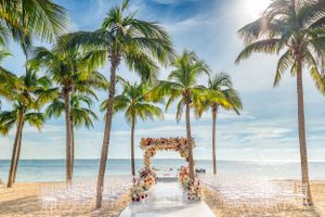 Tropical beach wedding setup with white chairs and floral arch between palm trees.