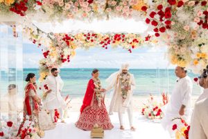 Beach wedding ceremony setup with floral arch and white chairs under red and pink flower decorations.