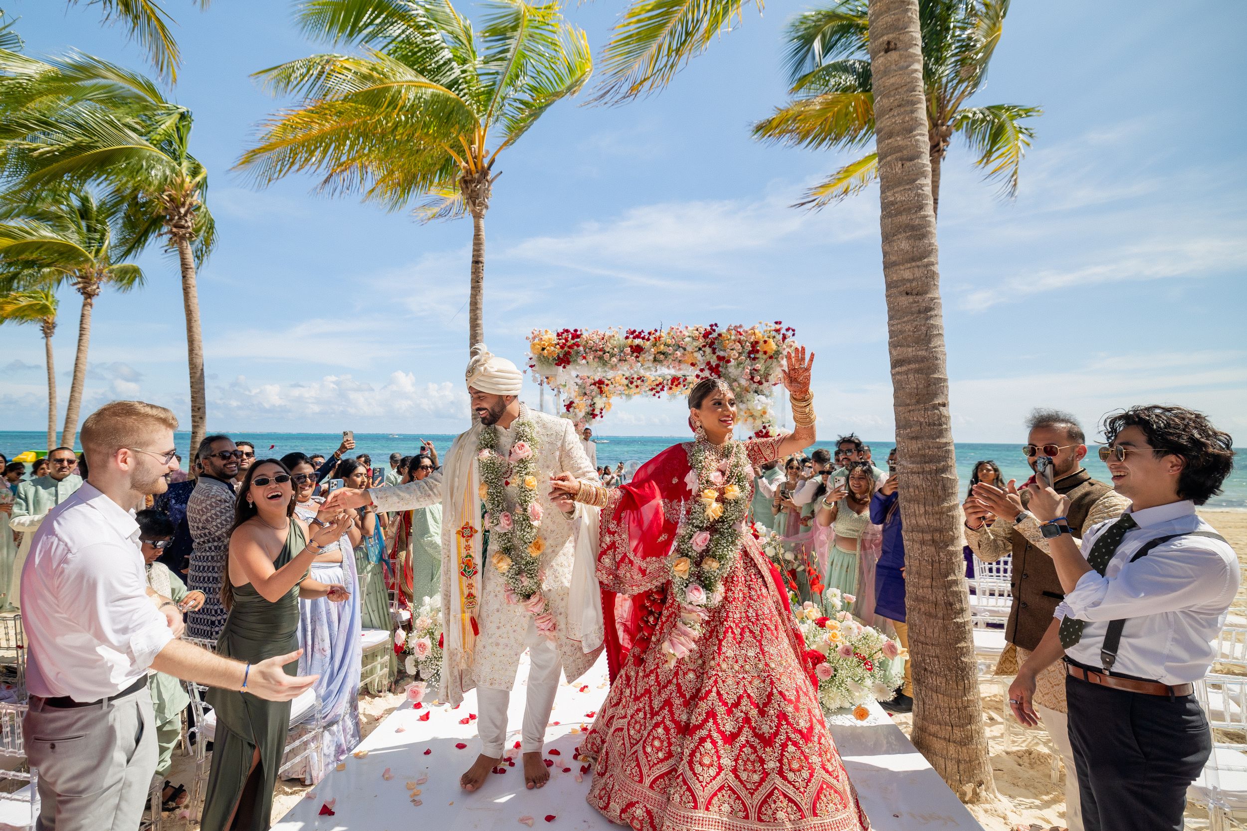 Outdoor beach wedding ceremony with guests gathered under floral arch and swaying palm trees.