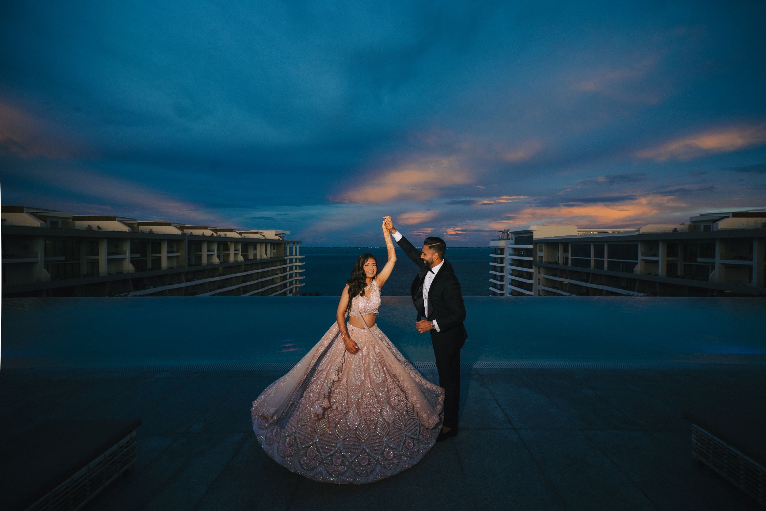 Couple in formal attire poses dramatically against dark blue sunset sky on rooftop.