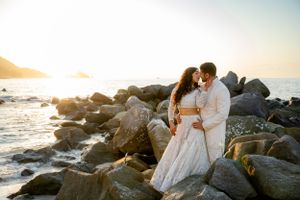 Couple in wedding attire embracing on rocky beach during golden hour.