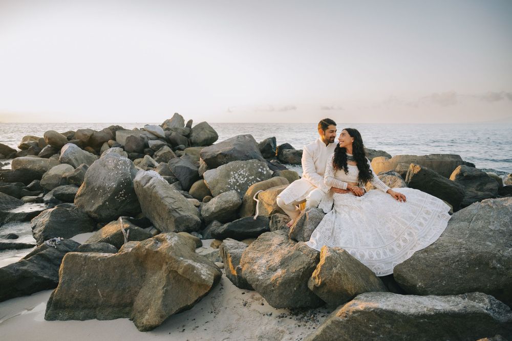 Romantic beach wedding photo with couple sitting on rocky coastline at sunset.