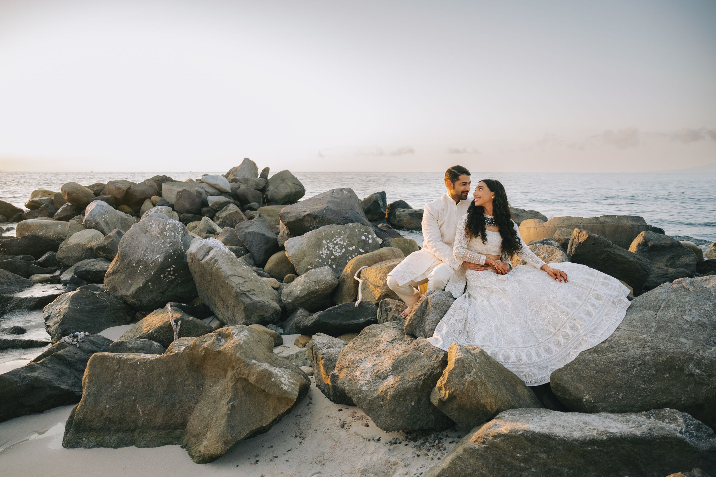 Romantic beach wedding photo with couple sitting on rocky coastline at sunset.