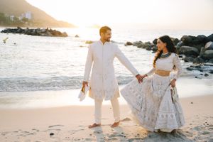 Couple in white attire walking hand in hand on beach at sunset.