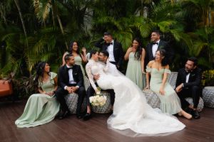 Wedding party in sage green bridesmaid dresses and black tuxedos posing together on tropical deck with palm trees.