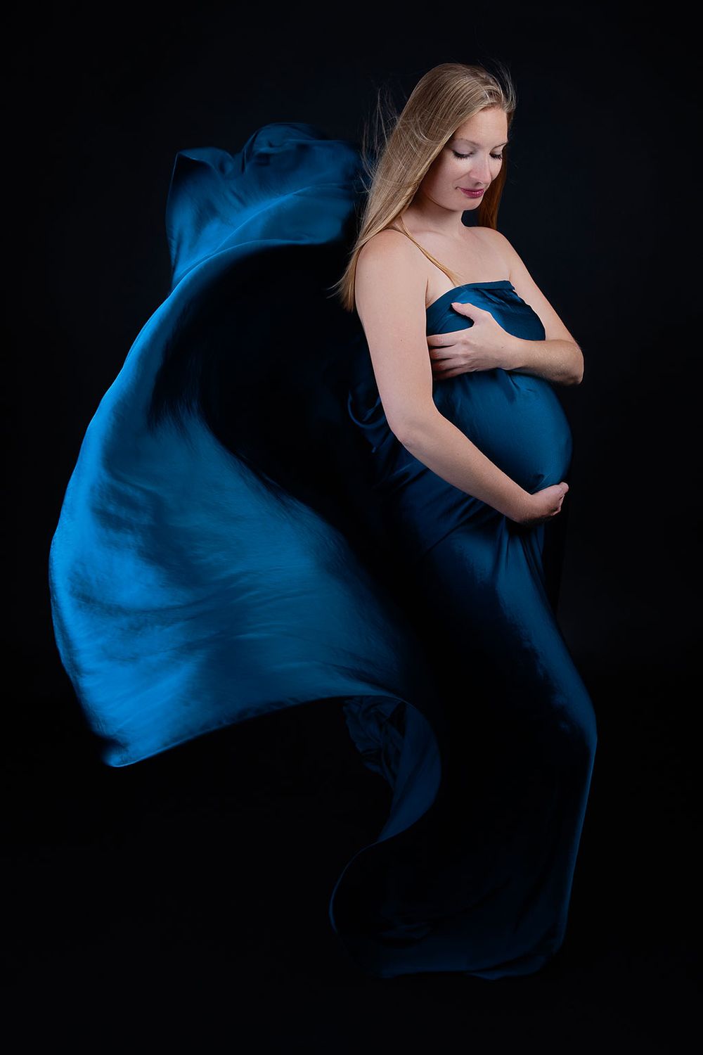 Elegant maternity portrait with billowing blue fabric on black backdrop.