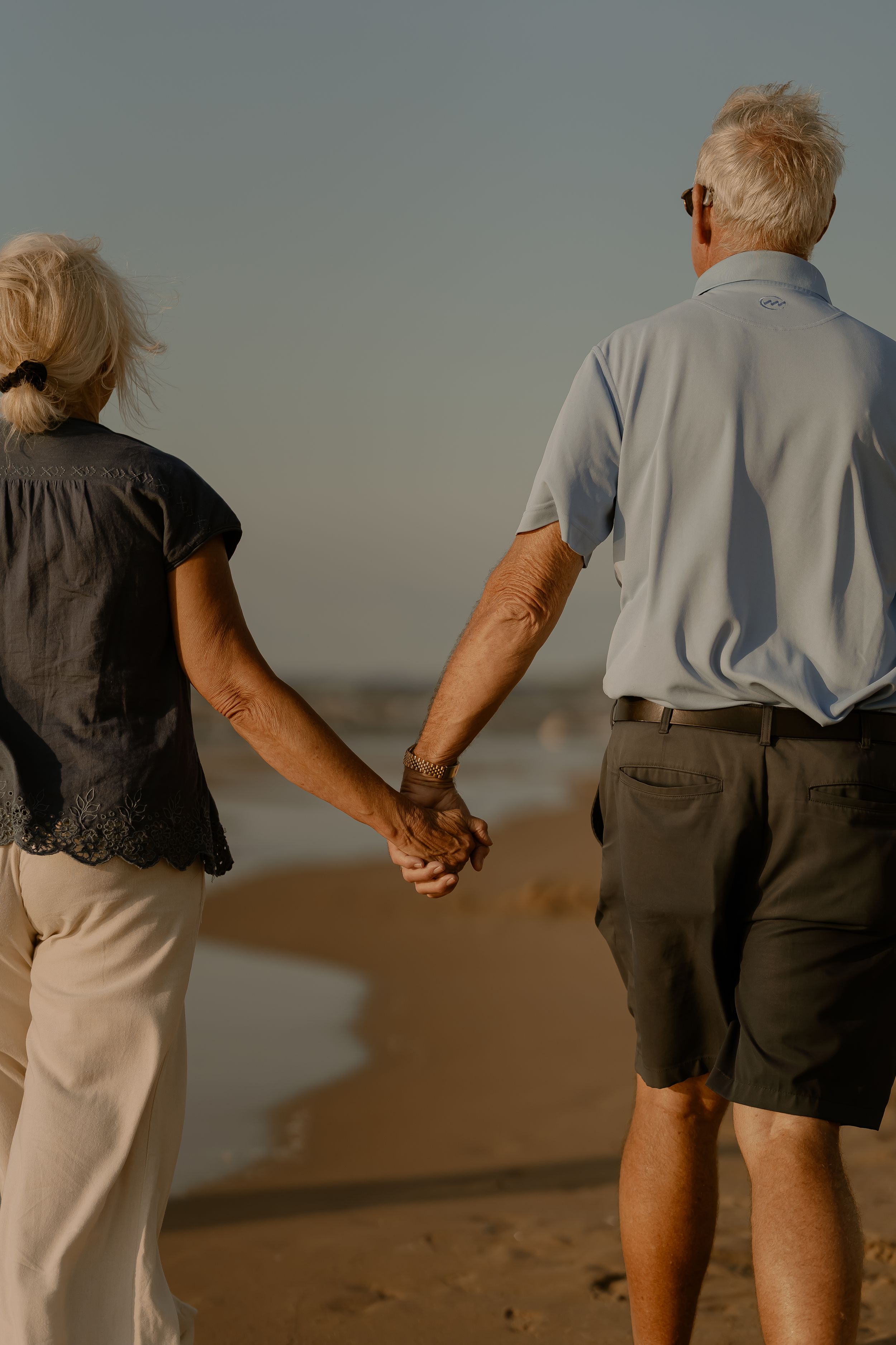 Elderly couple walking hand in hand along a sandy beach at sunset, photographed from behind.