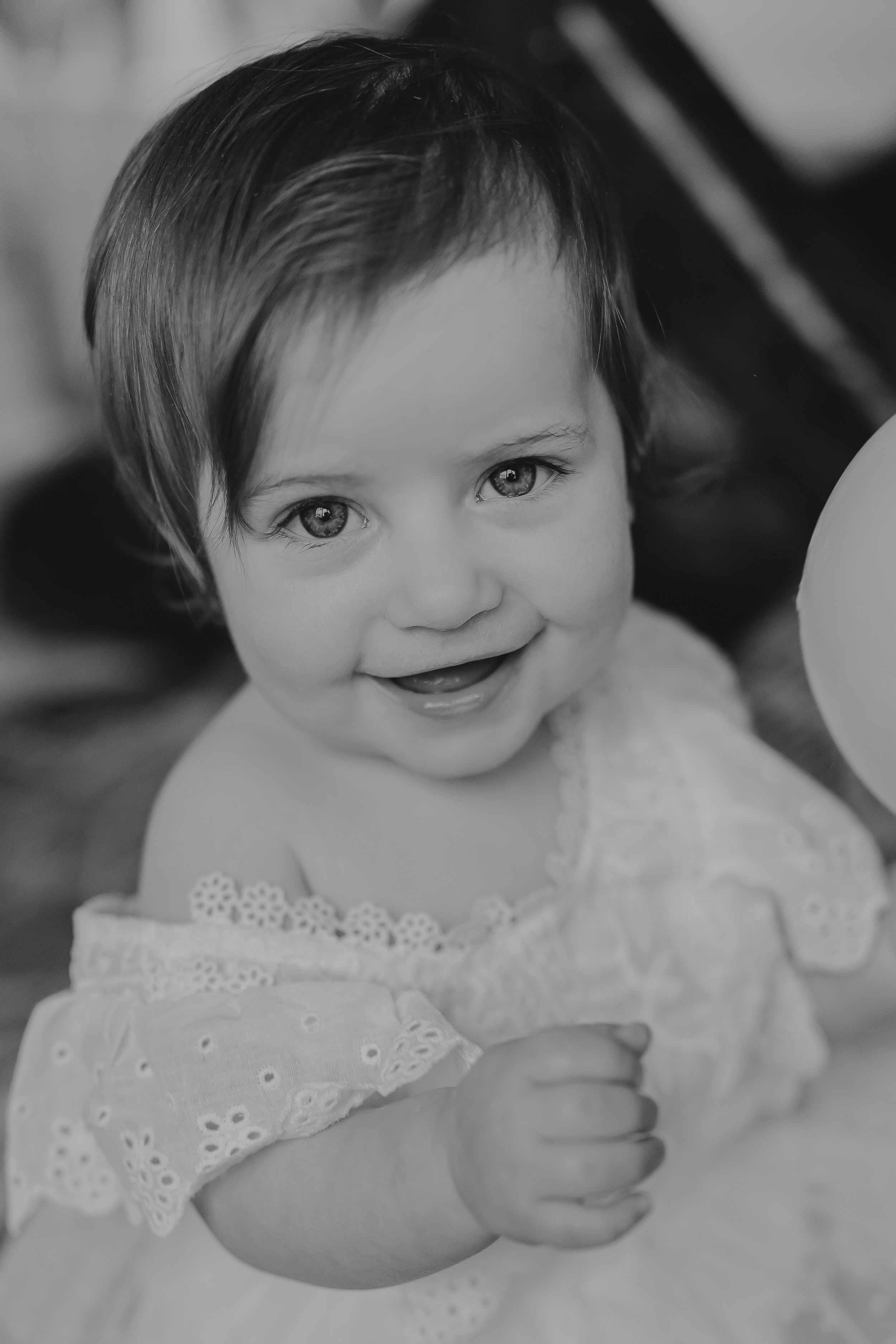 Black and white portrait of a smiling baby wearing a delicate white lace dress.