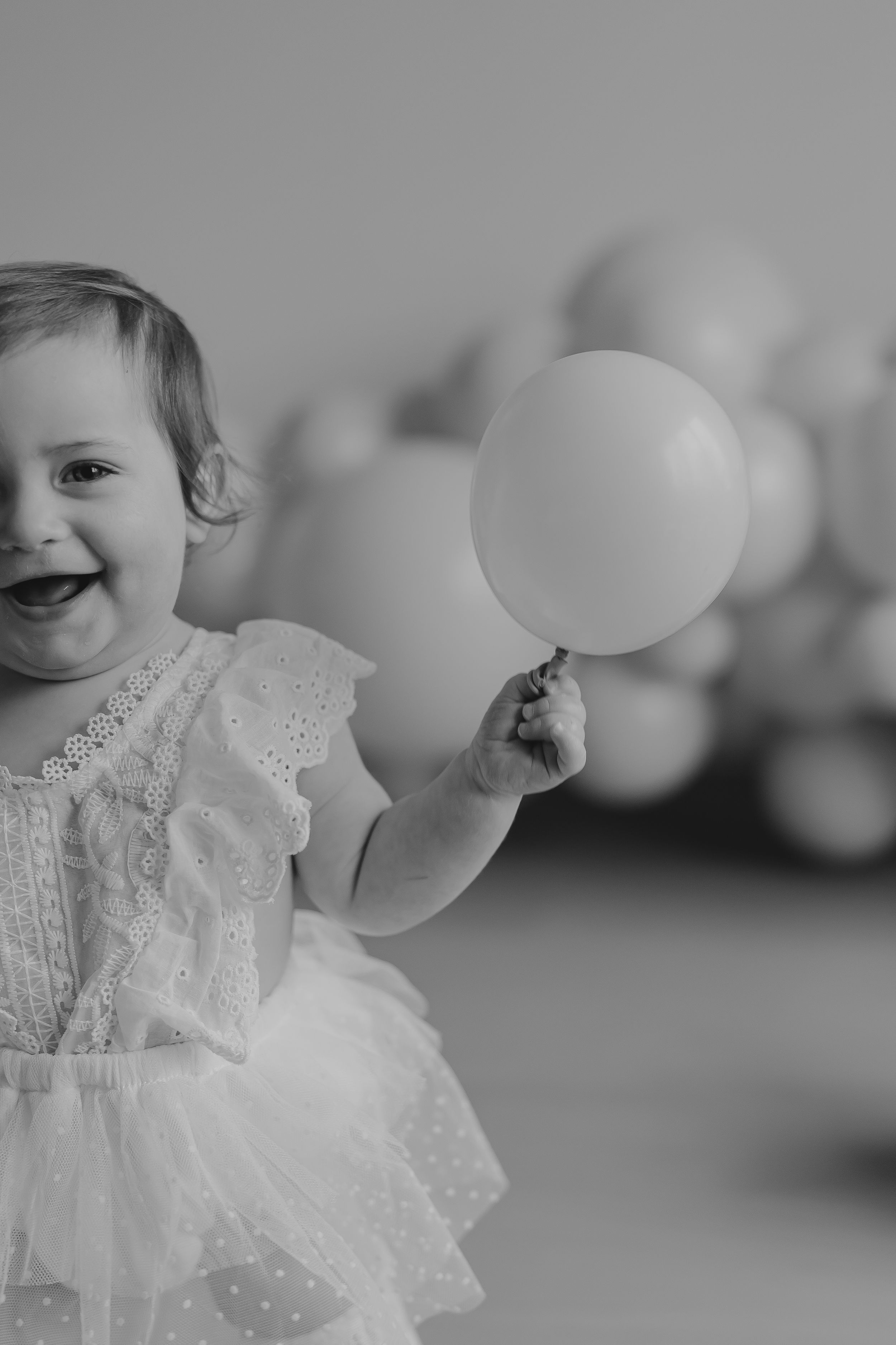 A baby in a white lace dress plays with white balloons in a black and white photograph.