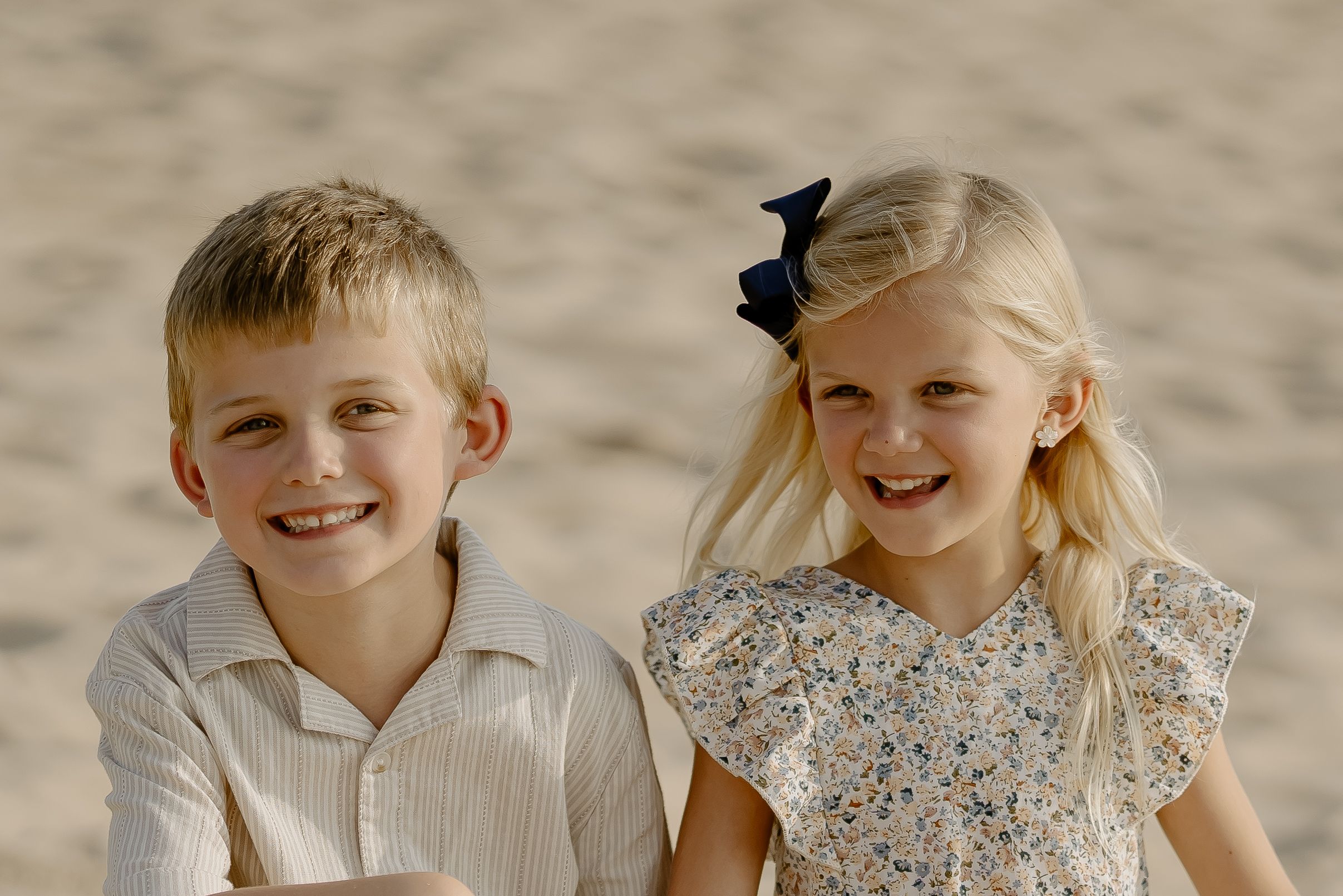Two young children sitting together outdoors on a sunny day, wearing light colored clothing.