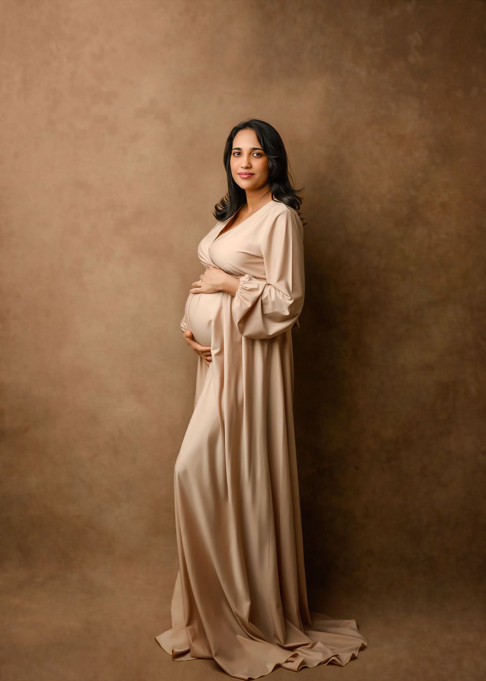 Elegant maternity portraits in flowing beige dress against brown studio backdrop.