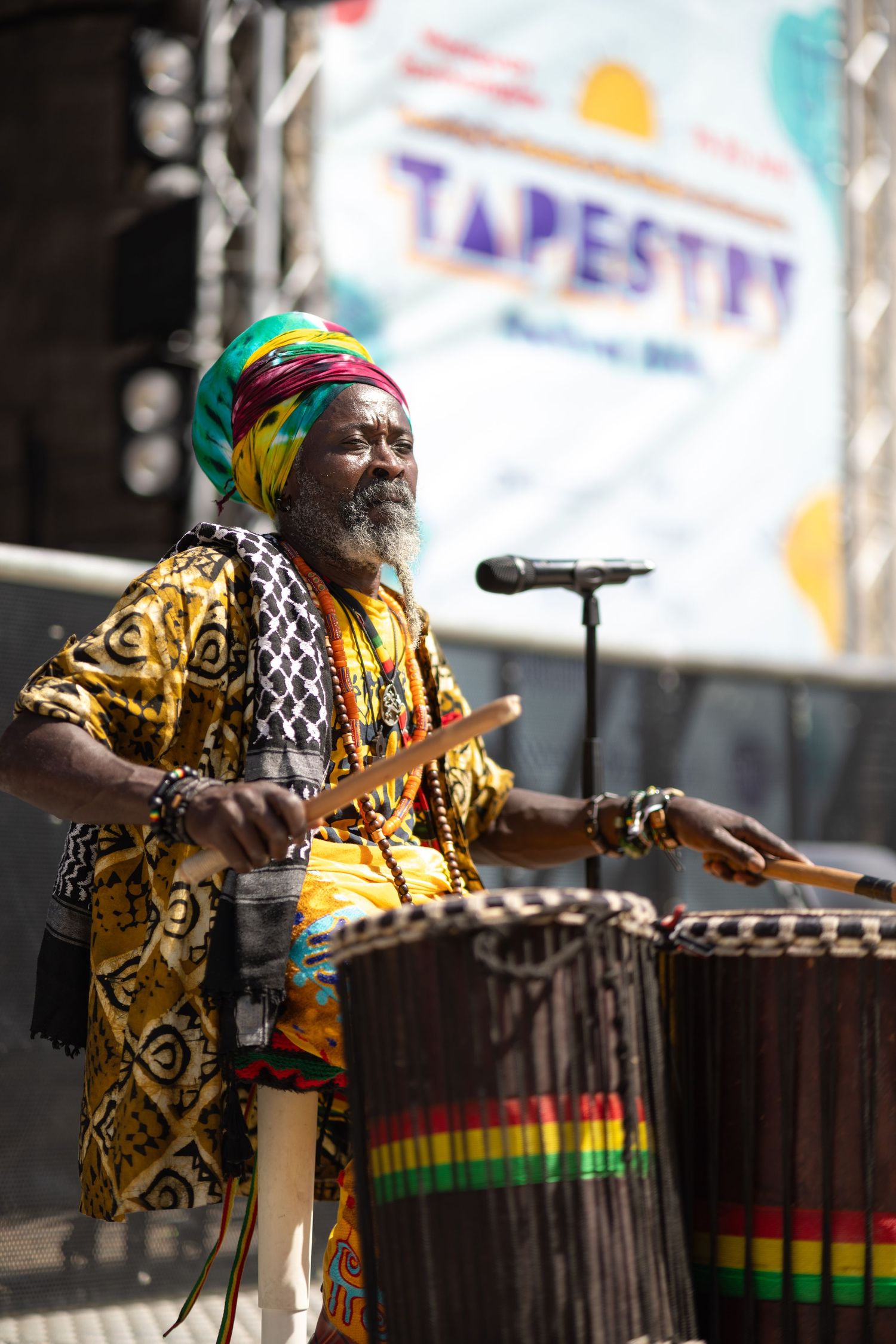 Musician in African clothing playing djembe drums at an outdoor festival with Tapestry banner, live music event photography