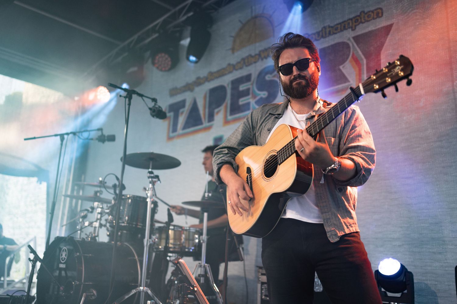 Musician in sunglasses playing acoustic guitar on stage at Tapestry festival with blue and red lighting, live music photography