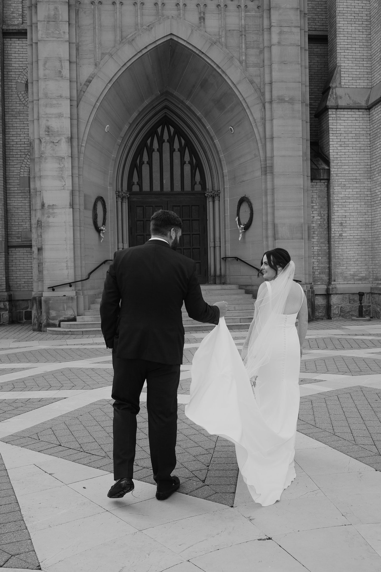Black and white photo of couple walking toward gothic church entrance with flowing wedding dress.