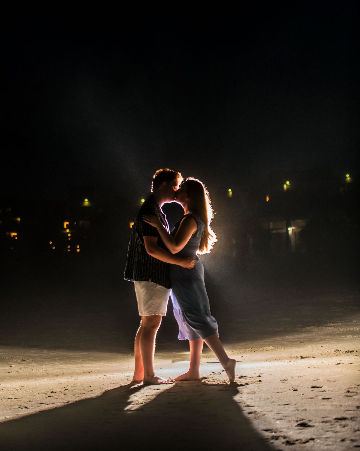 A silhouetted couple embracing on a dark beach with distant lights glowing. Myrtle beach photographer