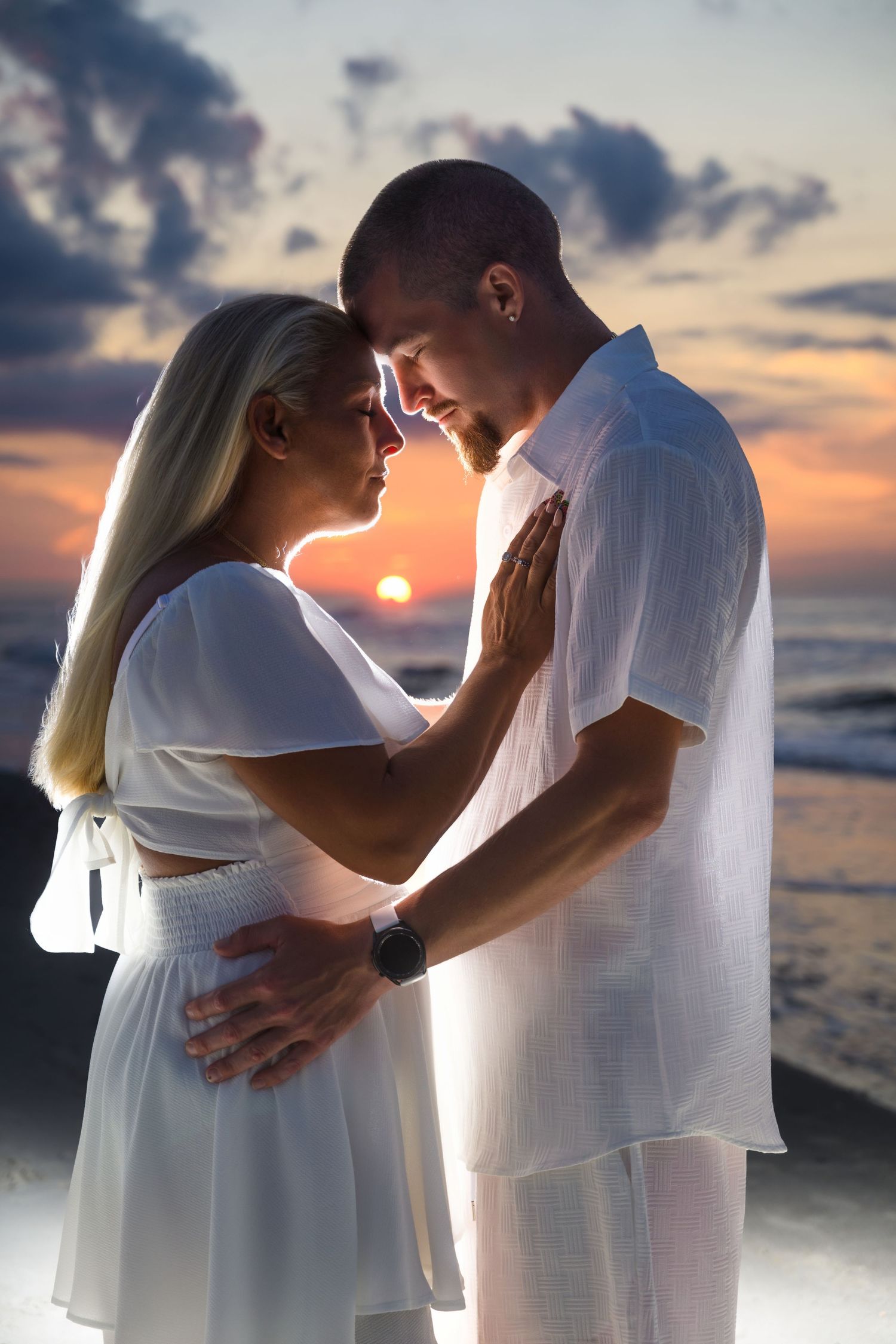A romantic couple in white clothing embrace on the beach at sunset while waves crash behind them. myrtle beach photographer