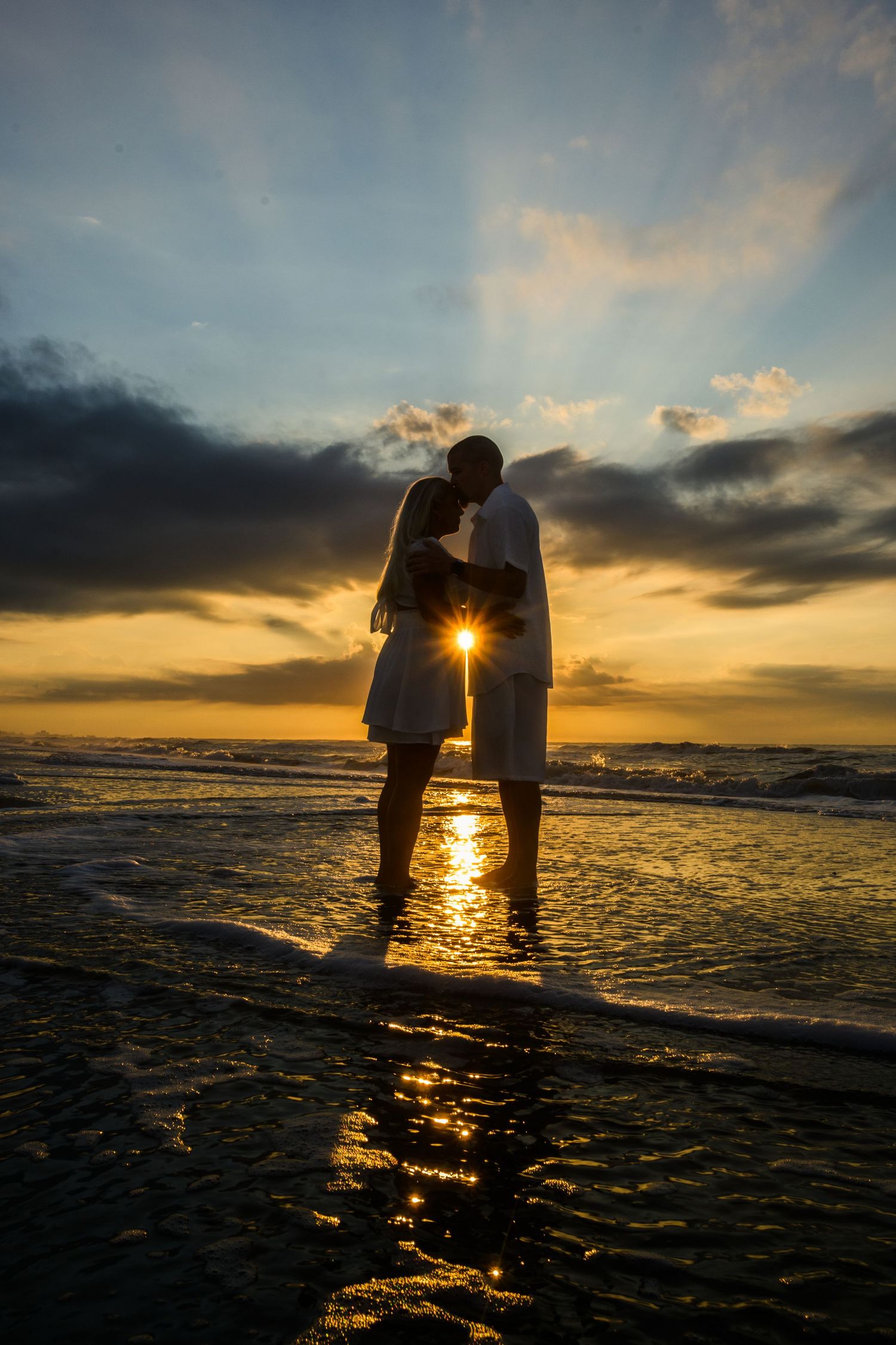 Silhouetted couple embraces on beach at sunset with golden light reflecting off ocean waves and dramatic sky. myrtle beach photographer