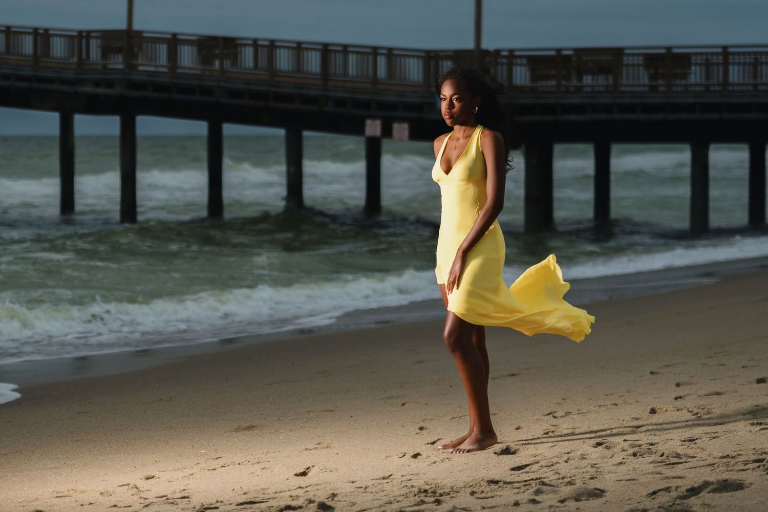 Woman in flowing yellow dress stands on beach near wooden pier at sunset. Myrtle beach photographer