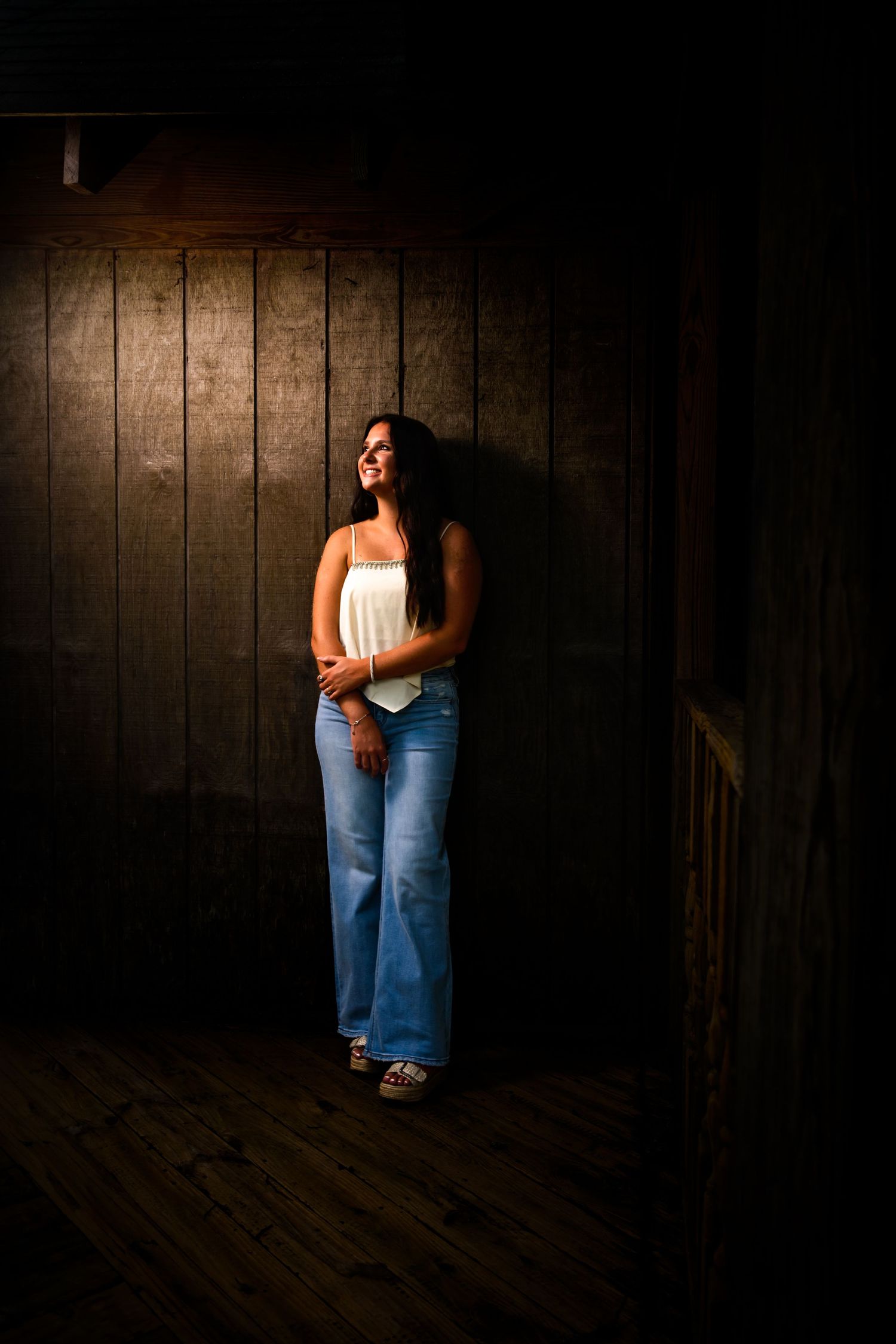 Woman in white top and flared jeans standing against dark wooden wall in dramatic lighting. Myrtle beach photographer