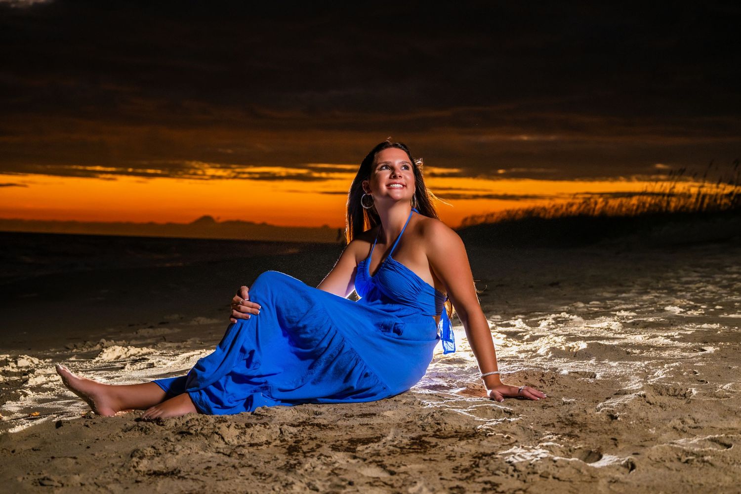 Figure in blue dress posed on sandy beach during dramatic orange sunset. Myrtle beach photographer
