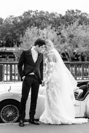 Black and white wedding portrait with vintage car against wooded background.