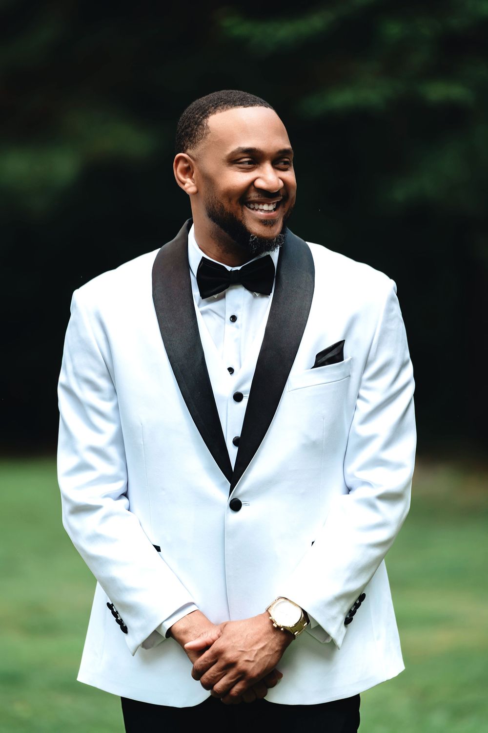 A stylish groom wearing a white and black tuxedo jacket with bow tie smiles joyfully in an outdoor setting.