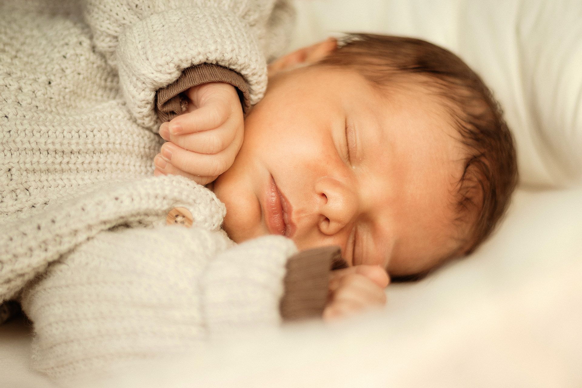 Ein schlafendes Neugeborenes im weißen Strickpullover liegt friedlich auf weißer Bettwäsche – Newborn Shooting Wiesbaden.