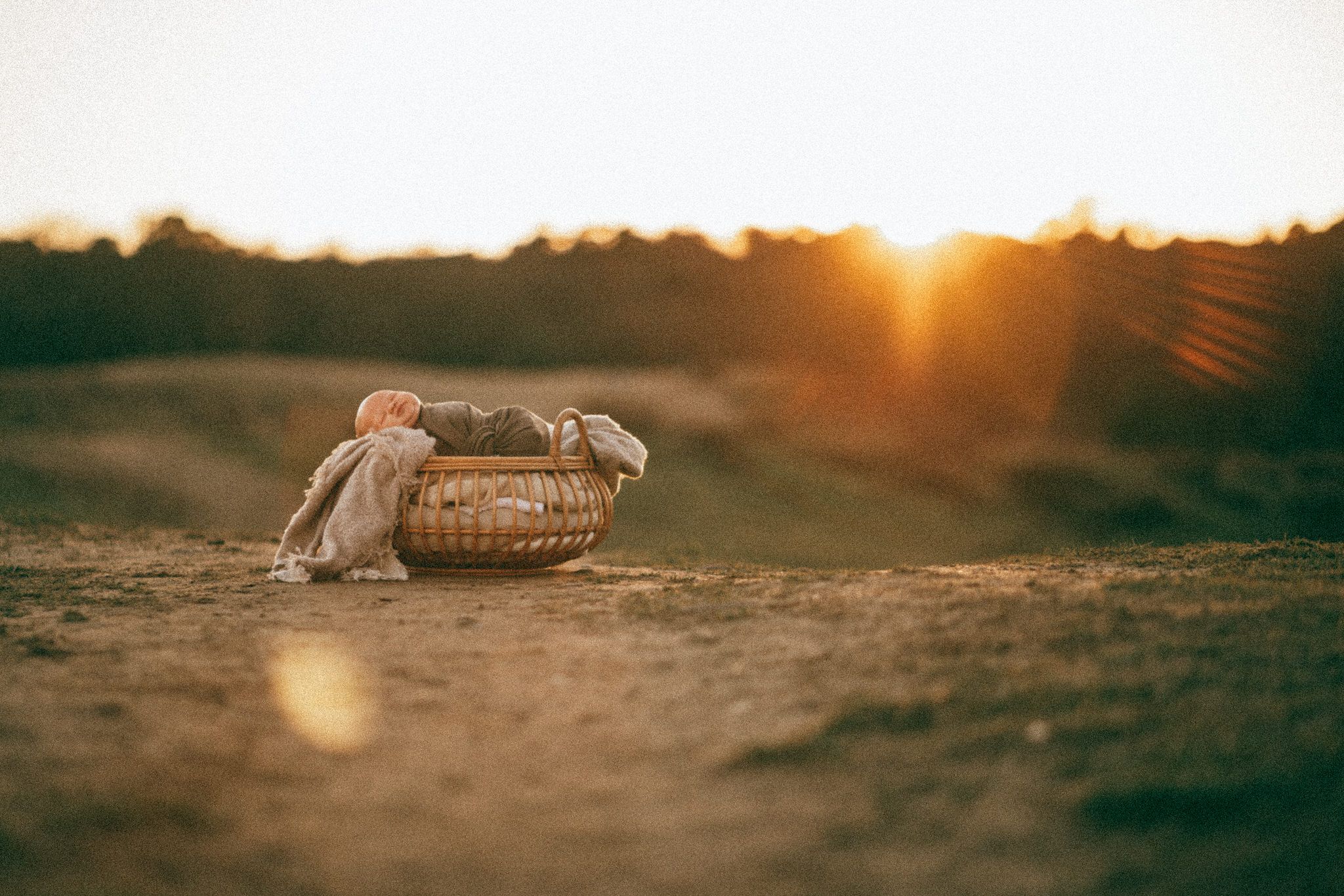 Ein Baby liegt in einem geflochtenen Korb mit Stoff, umgeben von staubigem Boden im goldenen Sonnenuntergang. Newborn Shooting Wiesbaden.
