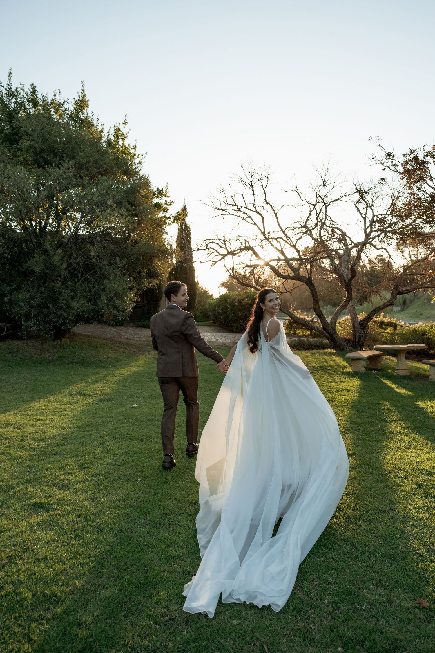 Bride in flowing white gown and groom in brown suit walk hand in hand through lush garden at golden hour.