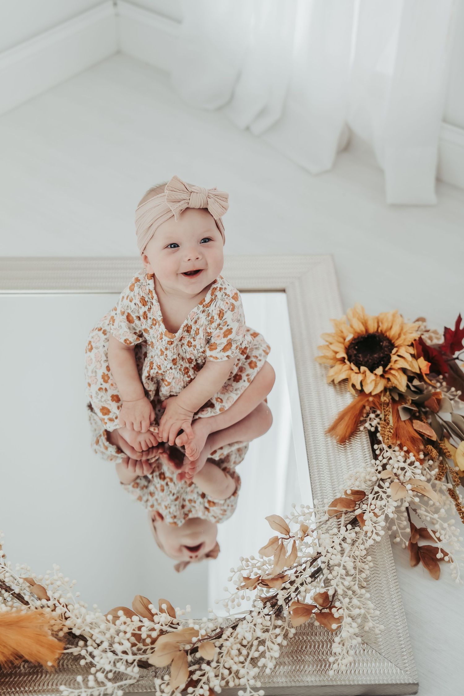 Baby wearing pearls and headband looking at reflection in mint green framed mirror on white floor.