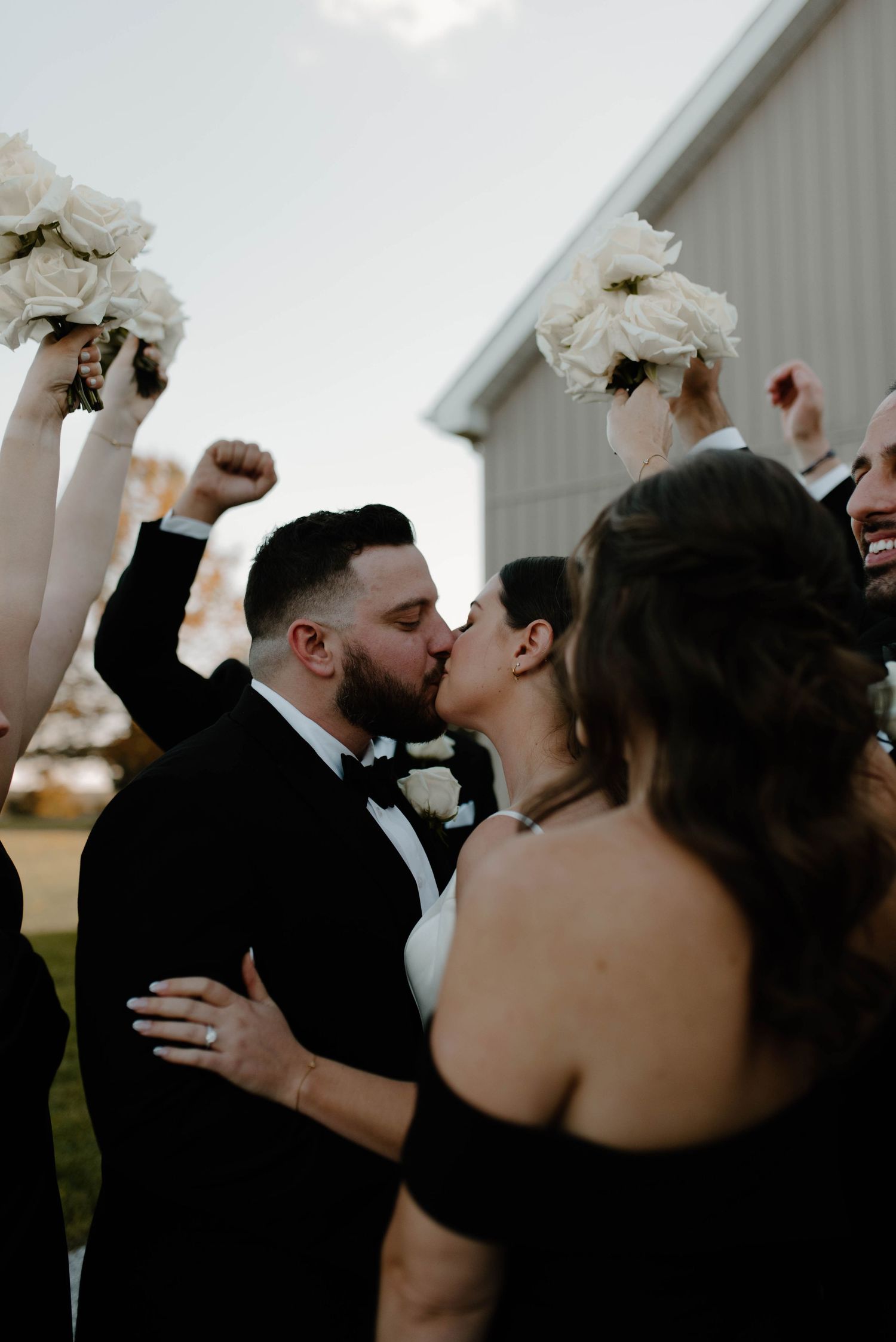 A newlywed couple kisses while wedding party members raise bouquets in celebration.