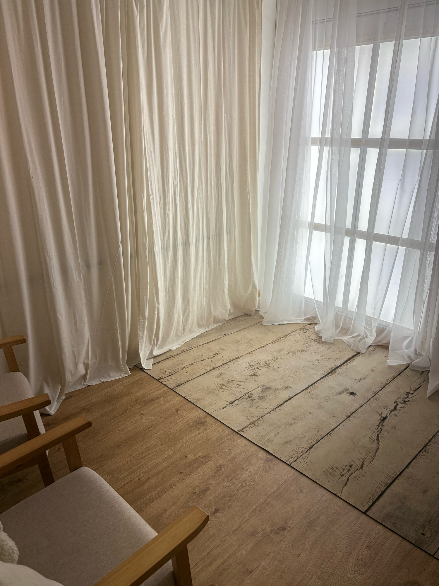 Bright room with white sheer curtains, wooden plank flooring, and wooden chairs in a minimalist interior setting.