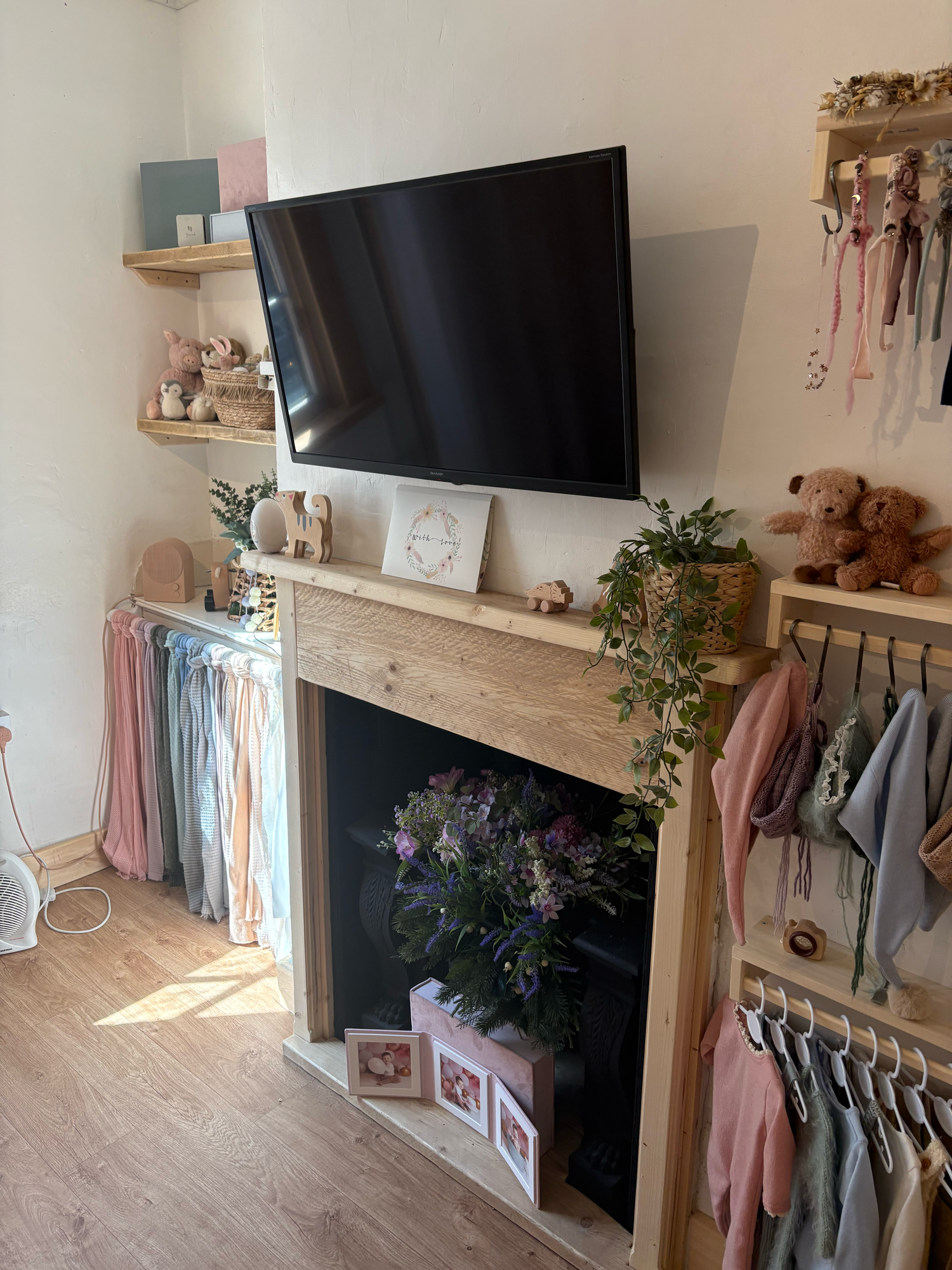 Cozy nursery room with wooden fireplace mantel, wall-mounted TV, floral arrangement, pastel garlands, and children's clothing display.
