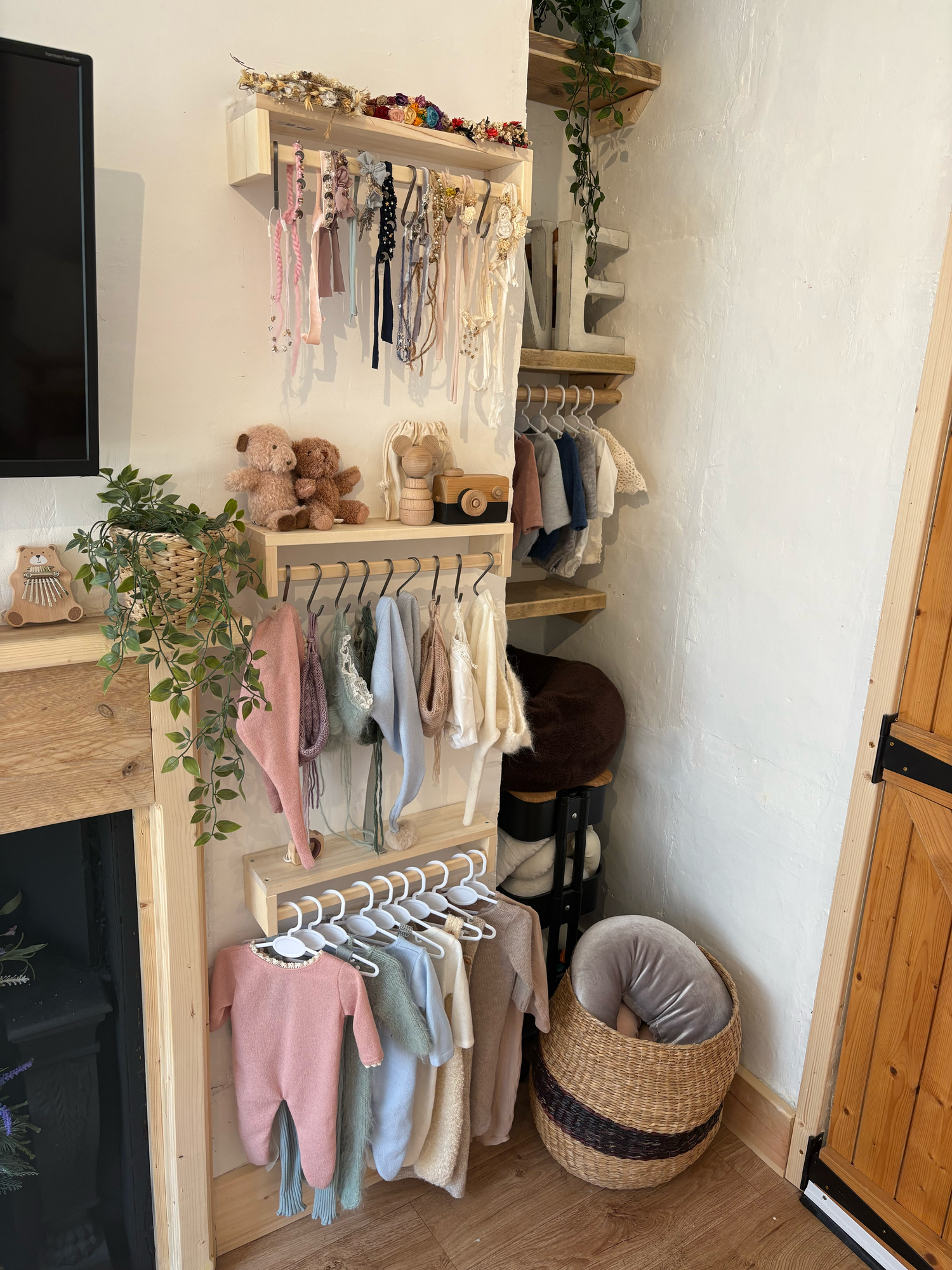Wooden nursery shelving unit displaying baby clothes on hangers, toys, and accessories with wicker baskets on hardwood floor.