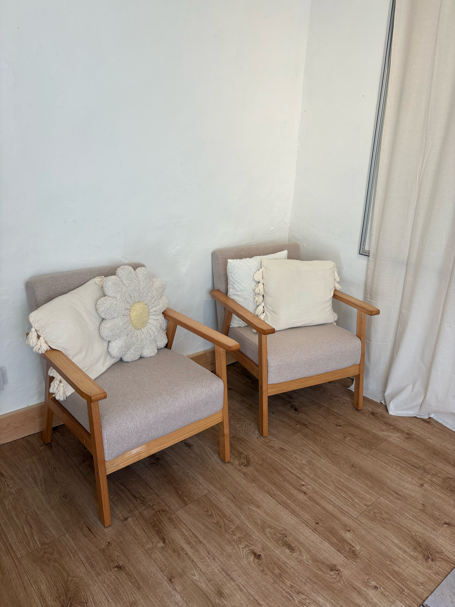 Two mid-century modern wooden armchairs with beige upholstery and decorative pillows on hardwood floor in corner.