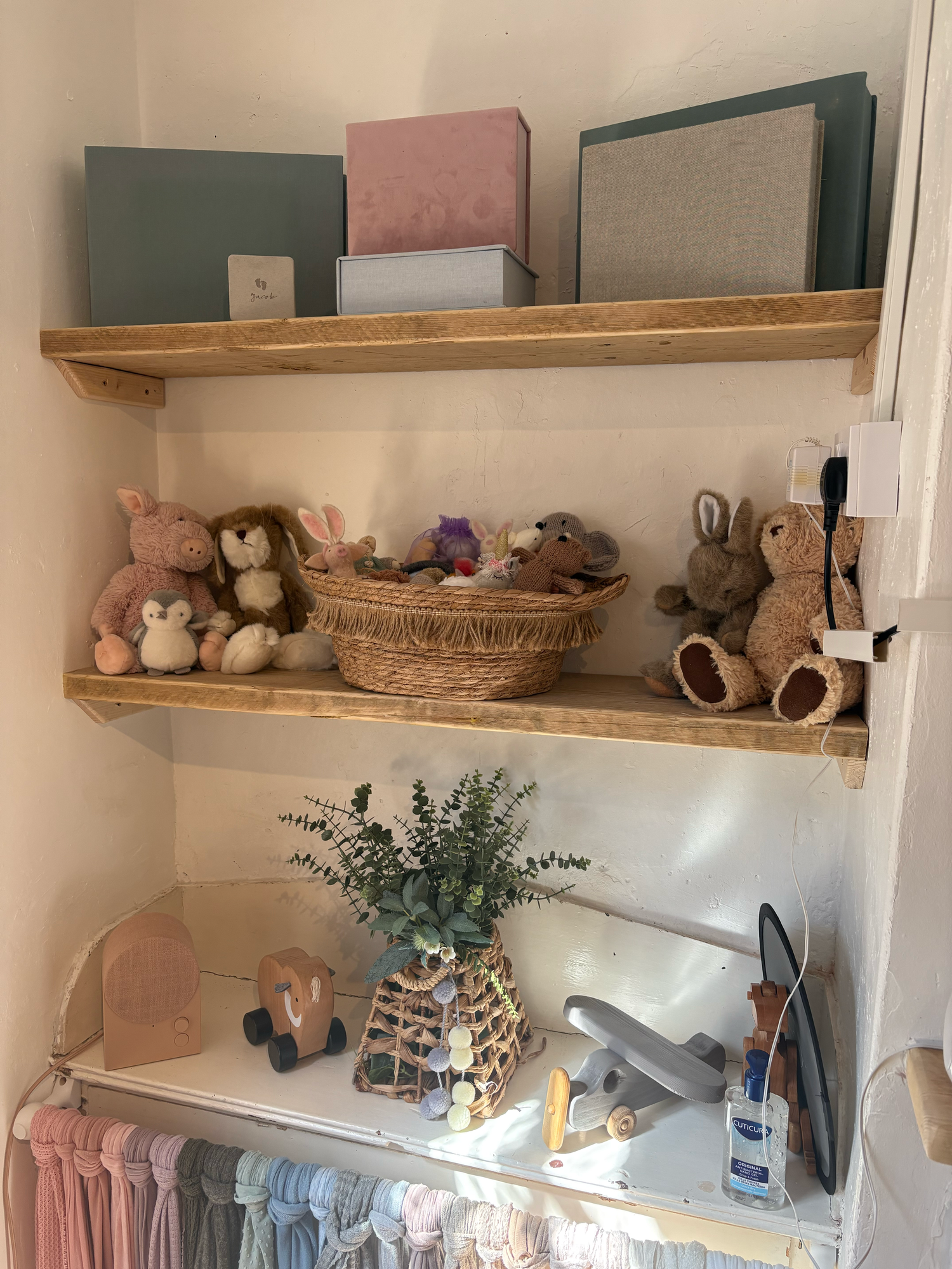 Wooden nursery shelves displaying stuffed animals, wicker basket, green boxes, and baby clothes on a hanging rail below.