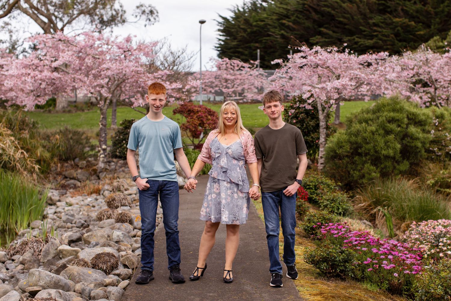 Mum and her sons standing together on a stone path beneath blooming pink cherry blossom trees in a garden at Palmerston North IPU.